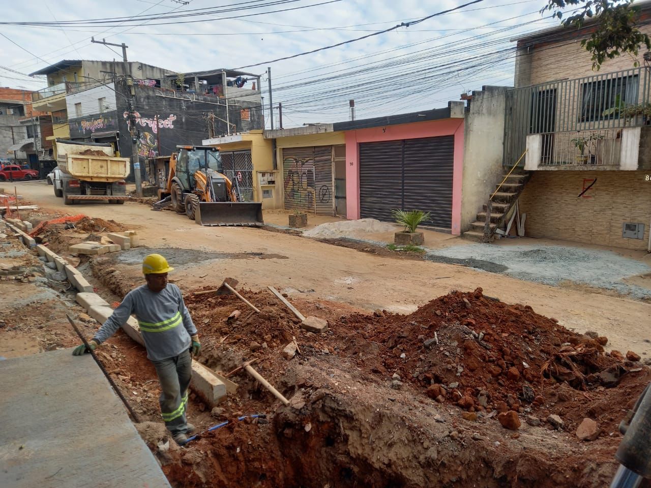Rua de terra, guias e sarjetas sendo construídas. Na imagem vemos um trabalhador passando, ele usa uniforme cinza com detalhes verdes e um capacete de segurança da cor amarela.
